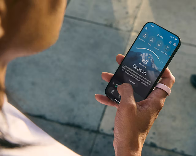A person views the Oura app on their smartphone while wearing a Petal Oura Ring Ceramic on their right index finger